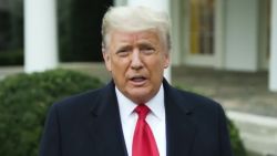 The image depicts former US President Donald Trump standing in front of the White House, wearing a red tie and suit.